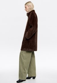 Abrigo de piel sintética marrón con cuello alto, bolsillos laterales y corte relajado, combinado con pantalones de pierna ancha verde claro y botas de tacón negro.