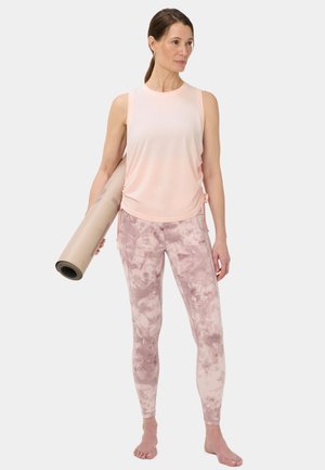 Femme en haut sans manches rose et leggings tie-dye tenant un tapis de yoga roulé, debout pieds nus devant un fond uni.