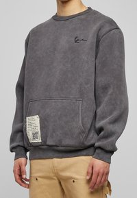 Šedá mikina vyrobená z bavlněné směsi, s přední klokankou, vyšitým logem a texturovanou nálepkou s tištěnými informacemi.