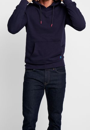 Marineblaue Kapuzenjacke mit einer Fronttasche, Kordelzugkapuze und rot akzentuierten Spitzen. Getragen mit dunklen Jeans. Weicher, elastischer Stoff.