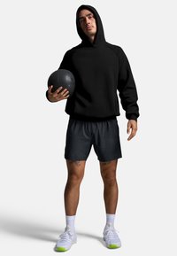 Melna kapucēta krekls, pelēki sporta īsie šorti, baltas sporta apavi ar zaļiem akcentiem, tur melnu vingrošanas bumbu. Sportisks piegriezums un ikdienišķs dizains.