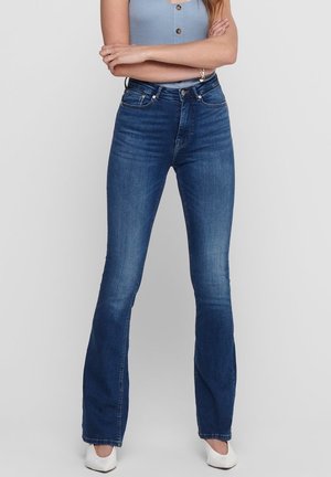 Femme portant un jean flare bleu foncé taille haute, un haut sans manches bleu clair, des chaussures blanches, debout les bras croisés devant un fond gris.