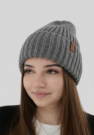 Gray knitted beanie with a ribbed texture and a brown leather tag. The hat is worn, covering the forehead and part of the hair.