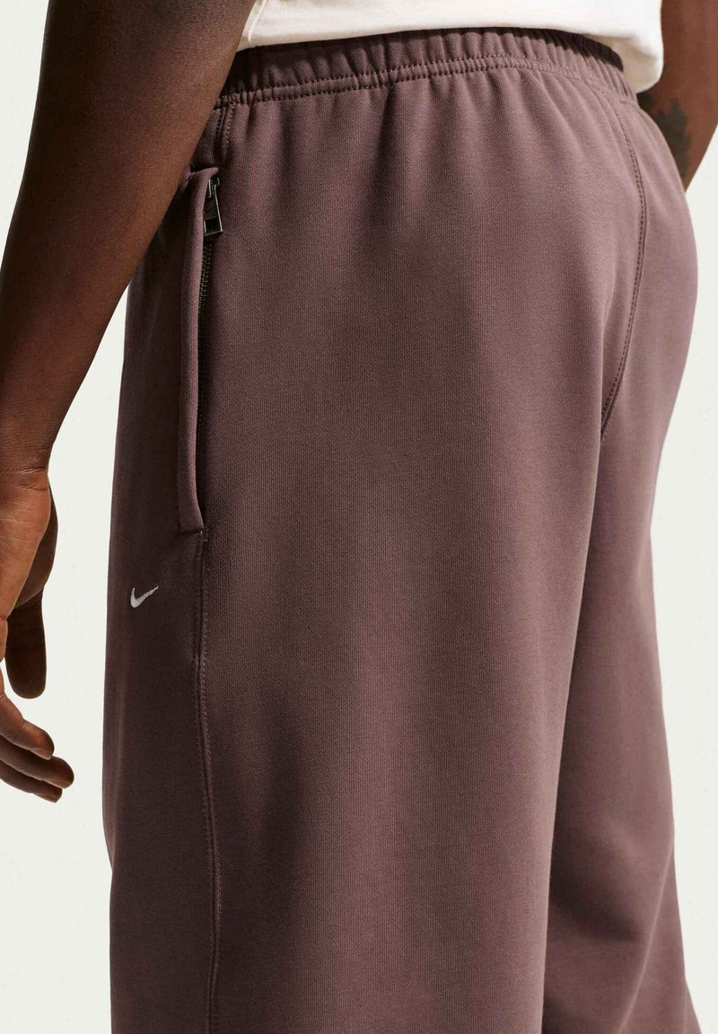 Persoon draagt mauve Nike-joggingbroek met een zak met rits, elastische tailleband en een wit shirt dat deels zichtbaar is bij de taille.