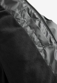 Chaqueta negra con un exterior de forro polar y un interior suave, con cierre de cremallera y un pequeño bolsillo en el interior.