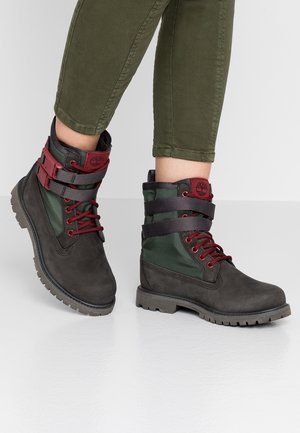 Bottes à lacets vert foncé et noir avec des accents bordeaux portées avec un pantalon vert olive retroussé sur un sol blanc.