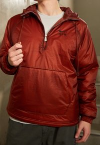 Röd lättviktsjacka med halvdragkedja, elastiska muddar och en framficka. Har en subtil ripstop-struktur och en logga på bröstet.