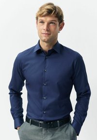 Sötétkék ing hosszú ujjal és hegyes gallérral, sötét gombokkal és sima, texturált anyagból készült. Szürke nadrággal párosítva.
