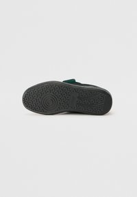 Suela de zapato de caucho negro con un patrón texturizado, detalles circulares y un acento verde en la parte superior. Presenta un diseño de perfil bajo.