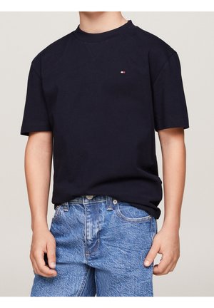 Marinblå bomullströja med rund hals, korta ärmar och en liten röd, vit och blå logotyp på bröstet; matchad med ljusblå denimjeans.