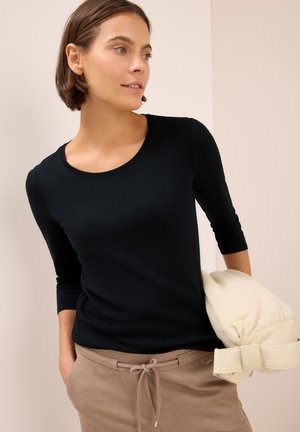 Top noir ajusté à manches longues avec un col rond, accompagné d'un pantalon beige et d'une veste rembourrée de couleur claire portée sur un bras.