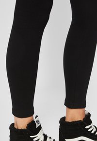 Svarta leggings och svarta högklackade sneakers med vita snören, som visar underbenen mot en ljus bakgrund.