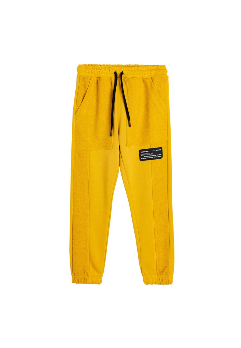 Gelbe Jogginghose mit elastischem Bund und Kordelzug, ausgestattet mit zwei Fronttaschen und einem schwarzen Etiketten-Detail. Weiche Stofftextur.