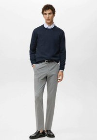 Man draagt marineblauwe trui over lichtblauw overhemd, grijze op maat gemaakte broek, zwarte riem en zwarte loafers, staand met handen gedeeltelijk in de zakken.