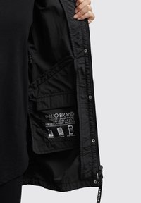 Main tenant une veste noire ouverte montrant une poche intérieure étiquetée pour passeport, smartphone, écouteurs, rouge à lèvres, chargeur portable et bouchons d'oreilles.