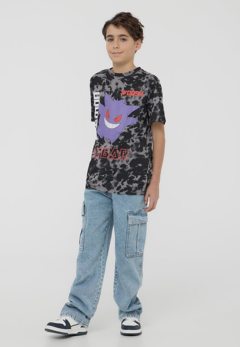 Maglietta tie-dye nera e grigia con un personaggio dei cartoni animati viola dagli occhi rossi, abbinata a pantaloni cargo blu leggermente usurati e scarpe da ginnastica.