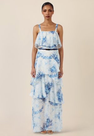 Frau trägt ein ärmelloses, bodenlanges weißes Kleid mit blauem Blumenmuster, gestuften Rüschen, dünnen Trägern und einem dünnen schwarzen Gürtel in der Taille.