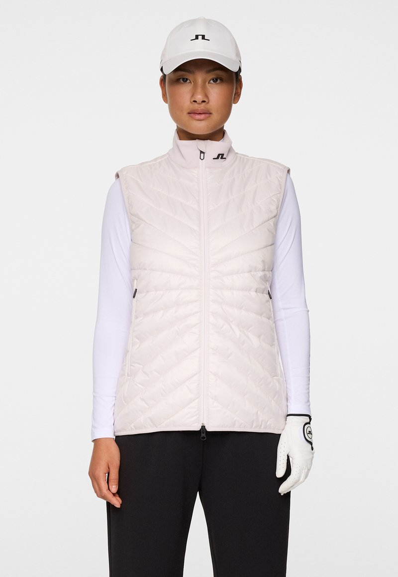 Ljusrosa, vadderad ärmlös väst med hög krage och framdragkedja, bärs över en vit långärmad skjorta, tillsammans med en keps och handskar.