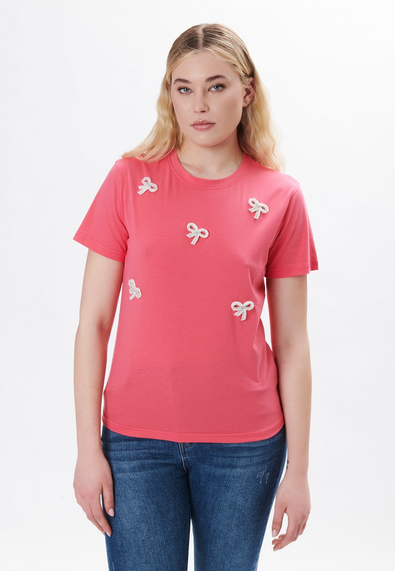 Koralfarvet t-shirt med rund udskæring, der har flere små hvide sløjfapplikationer. Stoffet virker blødt og let.