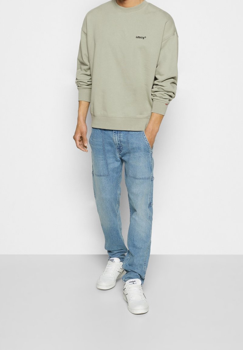 Homme portant un sweat-shirt Levi’s beige, un jean bleu clair avec poches à patch et des baskets blanches, debout avec une main dans la poche.