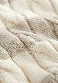 Strickdecke in Creme und Beige, mit strukturierten Zopfmustern und sanften Übergängen zwischen den Farbabschnitten. Weicher Wollstoff.