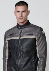 Crna i siva kožna moto jakna s patentnim zatvaračem sprijeda, stojećim ovratnikom i aplikacijama s logotipima brenda i riječi "RIDER."