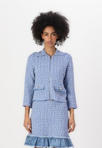 Blå tweedjakke med glidelås, to framlommer, og blå denimskjørt med volangkant. Begge har en strukturert veving og en tettsittende design.