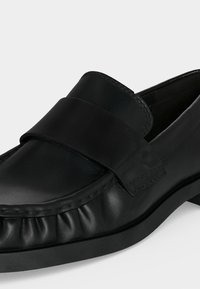 Μαύρος δερμάτινος μοκασίνι χωρίς κορδόνια με ραμμένη μύτη και φαρδιά λωρίδα πάνω από το ρομβοειδές μέρος, σε μαύρη λαστιχένια σόλα, παρουσιασμένο σε ελαφρώς πλάγια γωνία.