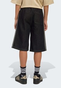 Shorts in denim nero con vestibilità leggermente ampia, caratterizzati da strisce laterali, una tasca posteriore e un orlo risvoltato. Indossati con calzini neri a righe e scarpe.