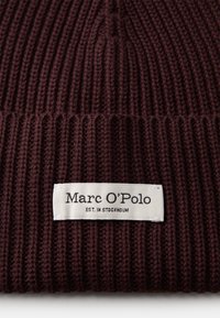 Gorro de punto acanalado marrón oscuro con un puño doblado que presenta una etiqueta rectangular blanca que dice "Marc O'Polo EST. EN ESTOCOLMO."