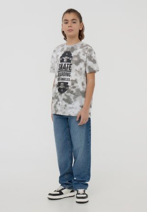 T-shirt grigia tie-dye con grafica di skateboard nera, abbinata a jeans blu rilassati e scarpe da ginnastica in bianco e nero. Design semplice e vestibilità casual.