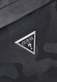 Juodas nailono piniginė su iškilniu raštu, turinti sidabrinę trikampę logotipo plokštelę. Lygi tekstūra su subtiliu blizgesiu ir aštriomis linijomis.