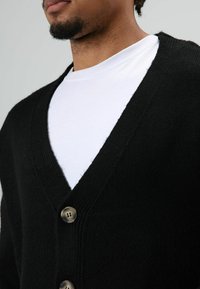 Cardigan noir à col en V, tissu texturé, avec trois gros boutons métalliques à l'avant, porté sur un T-shirt blanc.