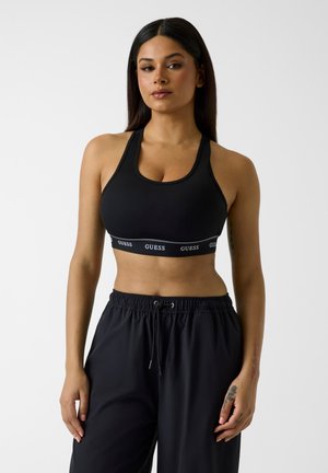 Femeie cu păr lung și închis la culoare, purtând un sutien sport negru cu bandă cu logo-ul "GUESS" și pantaloni largi negri cu șnur, stând în fața unui fundal alb.