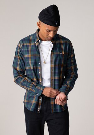 Mann justerer ermet på grønn og gul rutete knapp-opp-skjorte brukt over hvit t-skjorte, iført svart lue og svarte bukser.