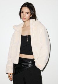 Jakna od kremaste umjetne kože s kapuljačom, patentnim zatvaračem sprijeda i trakama za stezanje. Nošena preko crnog kratkog topa i kožne suknje na preklop.
