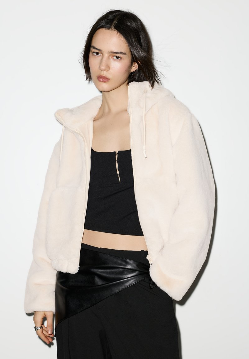 Kermaankermanvärinen tekoturkistakki, jossa huppu, vetoketju edessä ja kiristysnyöri. Päällä mustan cropatun toppiksen ja nahkaisen kietaisuhameen päällä.