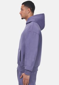 Lila Fleece-Hoodie mit Kängurutasche, gerippten Bündchen und Kapuze. Der Stoff wirkt weich mit einer glatten Textur.