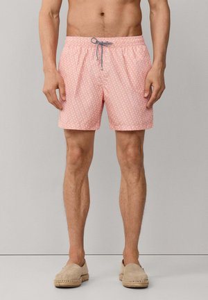 Homme portant un short de bain rose à motifs avec un cordon noir et blanc et des chaussures beiges à enfiler, se tenant devant un fond uni.