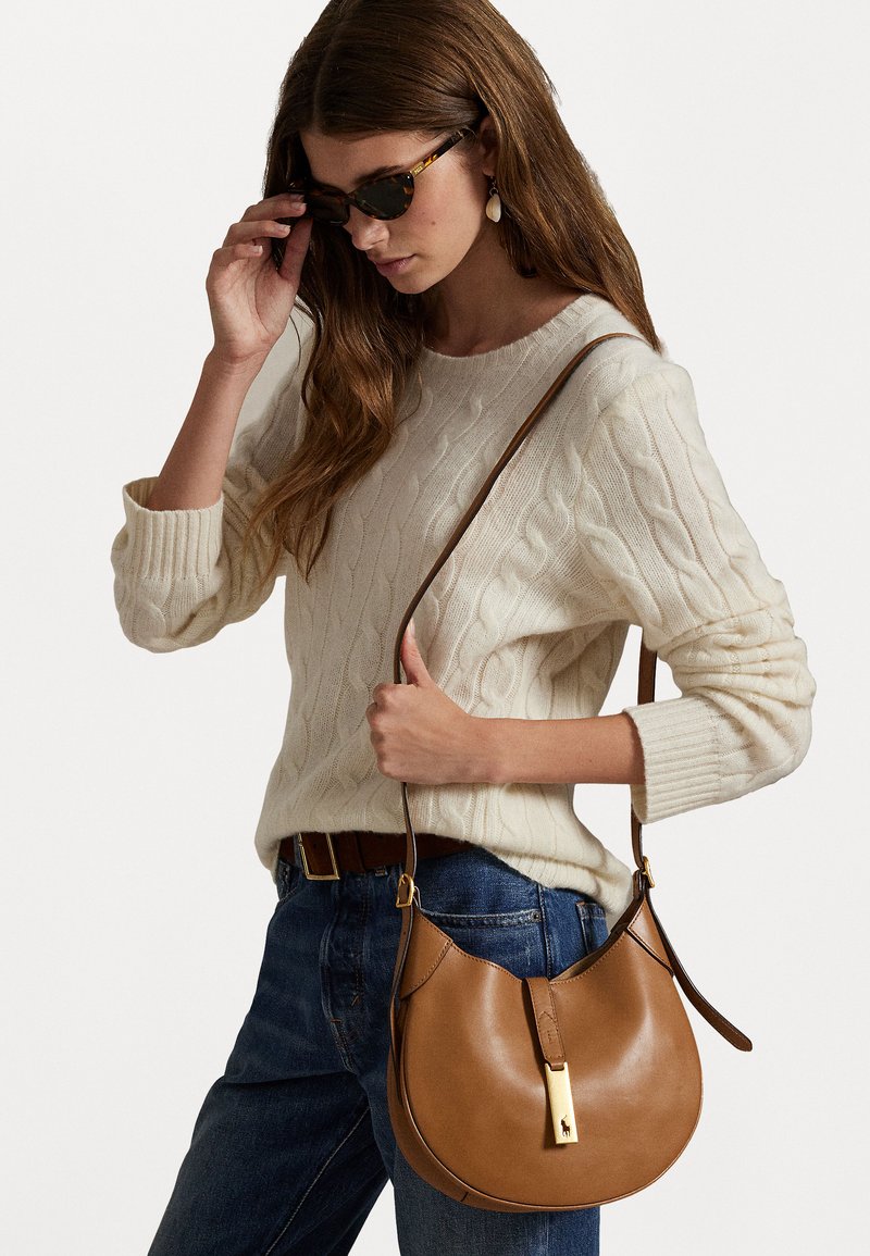 Bolsa de ombro castanha em couro com um formato curvo e detalhes em metal dourado. O modelo veste um suéter de malha com padrão de corda em tom creme e jeans azuis.