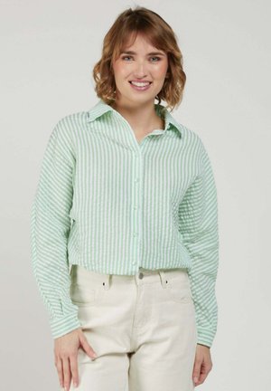 Jeune femme souriante, portant une chemise à rayures vert clair et blanc avec boutons et un pantalon couleur crème, debout devant un fond uni.