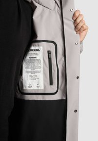 Gai pelēks žakets ar melniem akcentiem, ar rāvējslēdzēja iekšējo kabatu un etiķetes detaļām. Izgatavots no gludas, izturīgas materiāla.