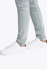 Λευκά αθλητικά παπούτσια με λείο δερμάτινο πάνω μέρος, στρογγυλεμένη μύτη και επίπεδη δέση με κορδόνια· διαθέτουν ναυτικό μπλε λεπτομέρεια στη φτ έρνα και υφή από καουτσούκ στην εξωτερική σόλα.