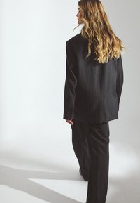 Black pinstriped suit with oversized silhouette; structured blazer and tailored trousers. Wavy hair visible, light background.