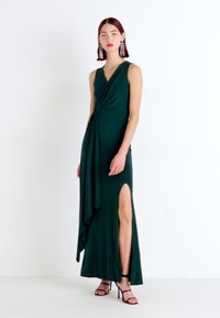 Rochie maxi fără mâneci, de un verde smarald, cu detalii drapate și fanta laterală; stofă netedă și siluetă fluidă. Asortată cu sandale negre cu curele.