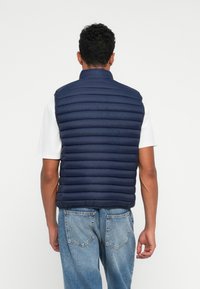 Marineblå puffervest med vandret quilted tekstur, høj krave og uden ærmer. Båret over en hvid t-shirt og lyseblå jeans.