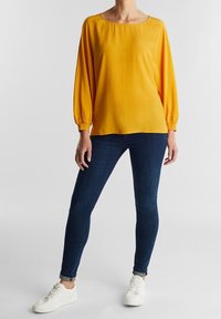 Haut jaune léger à manches longues avec un col rond, associé à un jean ajusté bleu foncé et des baskets blanches. Texture lisse.