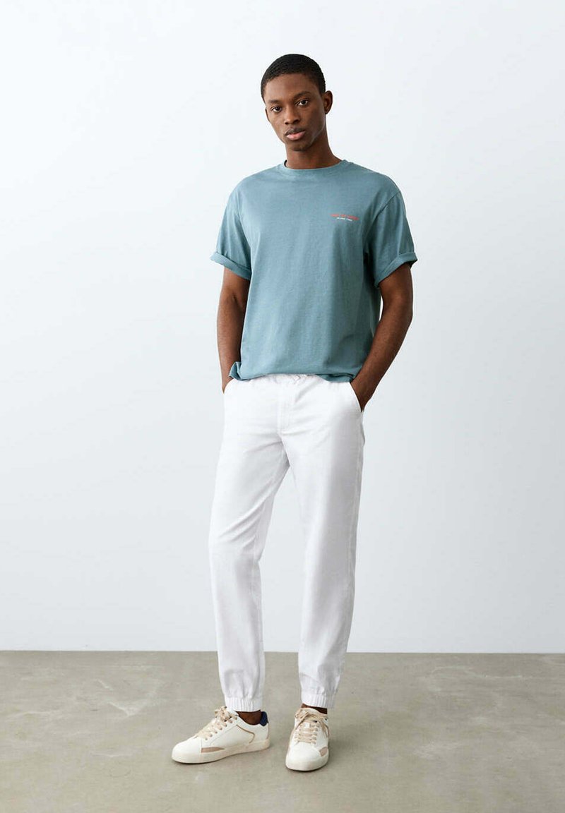Jeune homme debout, les mains dans les poches d’un pantalon jogger blanc, portant un t-shirt bleu sarcelle à manches retroussées et des baskets blanches sur un sol gris.