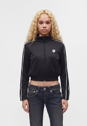 Jeune femme aux cheveux blonds bouclés portant une veste noire zippée avec des rayures blanches et un jean foncé, debout devant un fond clair uni.