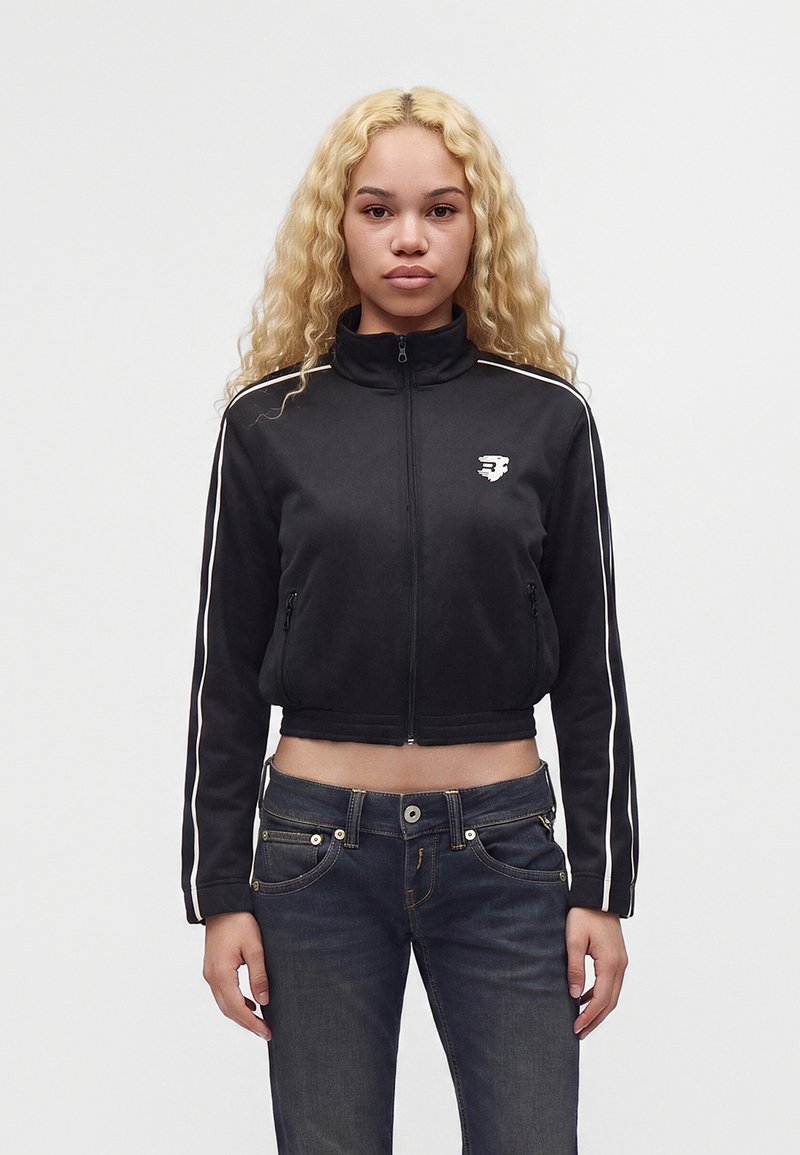 Jeune femme aux cheveux blonds bouclés portant une veste noire zippée avec des rayures blanches et un jean foncé, debout devant un fond clair uni.